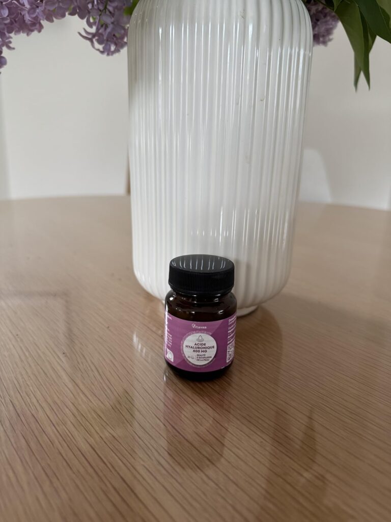 Le flacon d'acide hyaluronique posé sur une table en bois, devant un vase blanc et un bouquet de fleurs violettes. Le flacon...