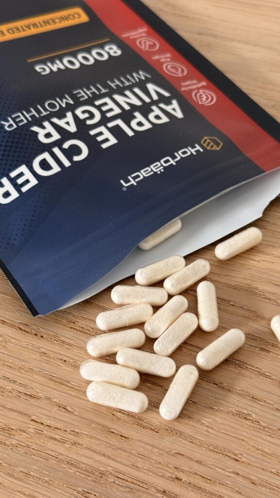 Vue rapprochée des capsules de compléments alimentaires Horbaach, légèrement dispersées sur une surface en bois claire. Le sa...