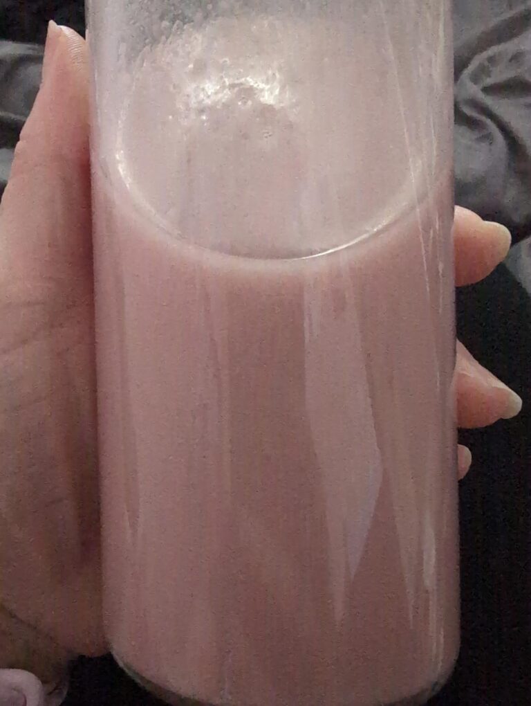 Une main tenant un verre transparent rempli du smoothie protéiné Kiba. Le liquide est de couleur rose pâle, et l'arrière-plan...
