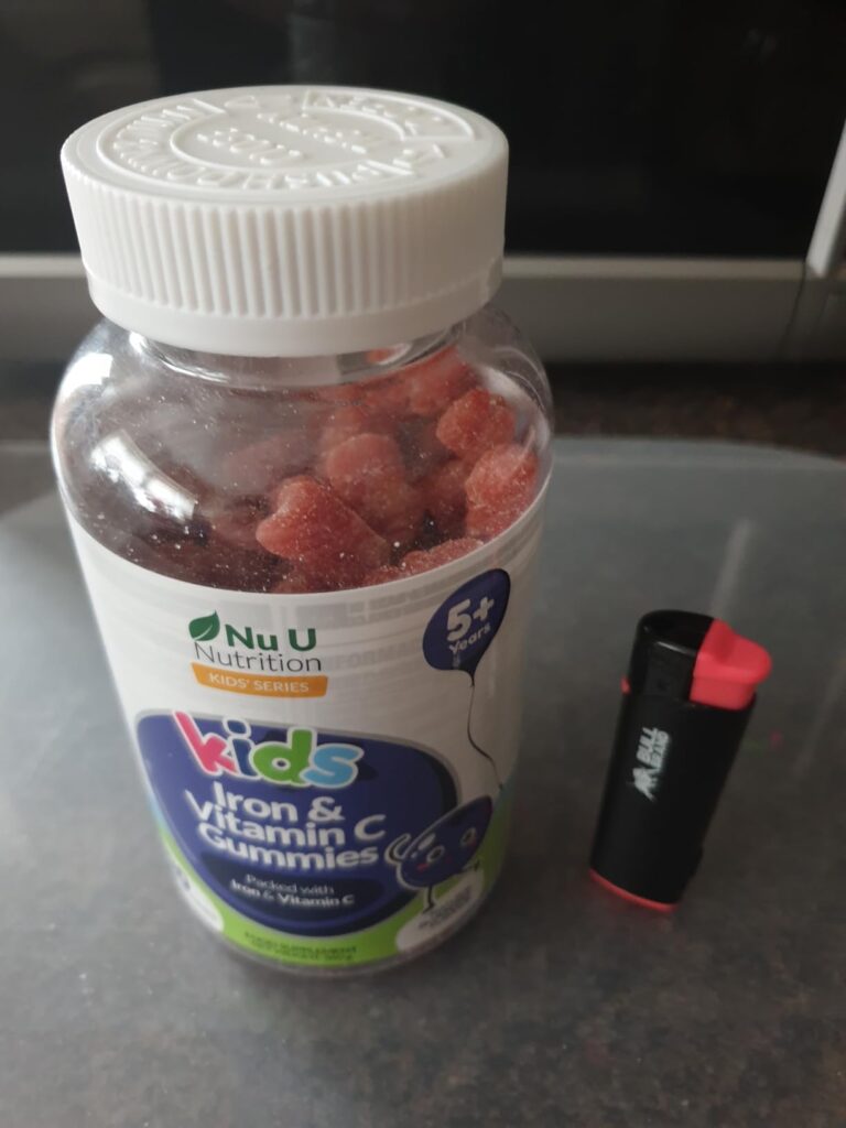 Bouteille de vitamines pour enfants, posée sur une table, avec un briquet à côté pour donner une idée de la taille.