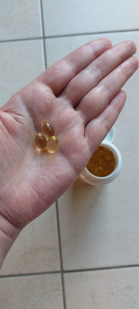 Une main tient trois capsules de compléments alimentaires, tandis que le flacon est visible en arrière-plan avec d'autres cap...