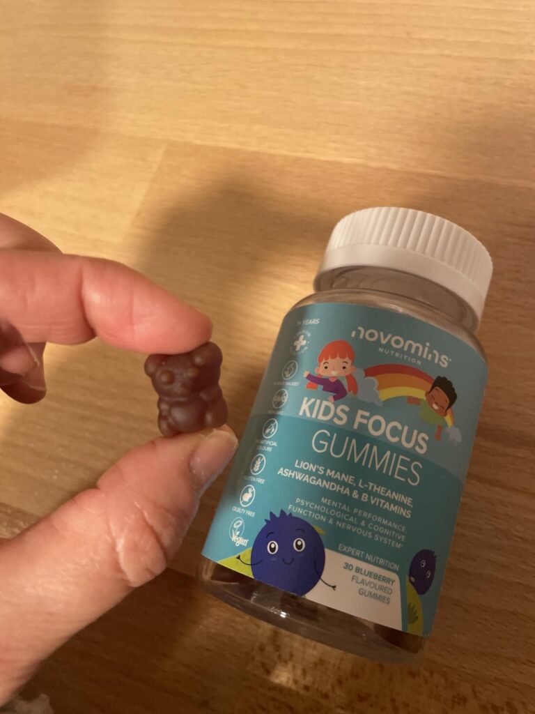 Une main tenant une gomme de forme d'ours marron, avec le flacon de Novomins Kids Focus Gummies en arrière-plan.