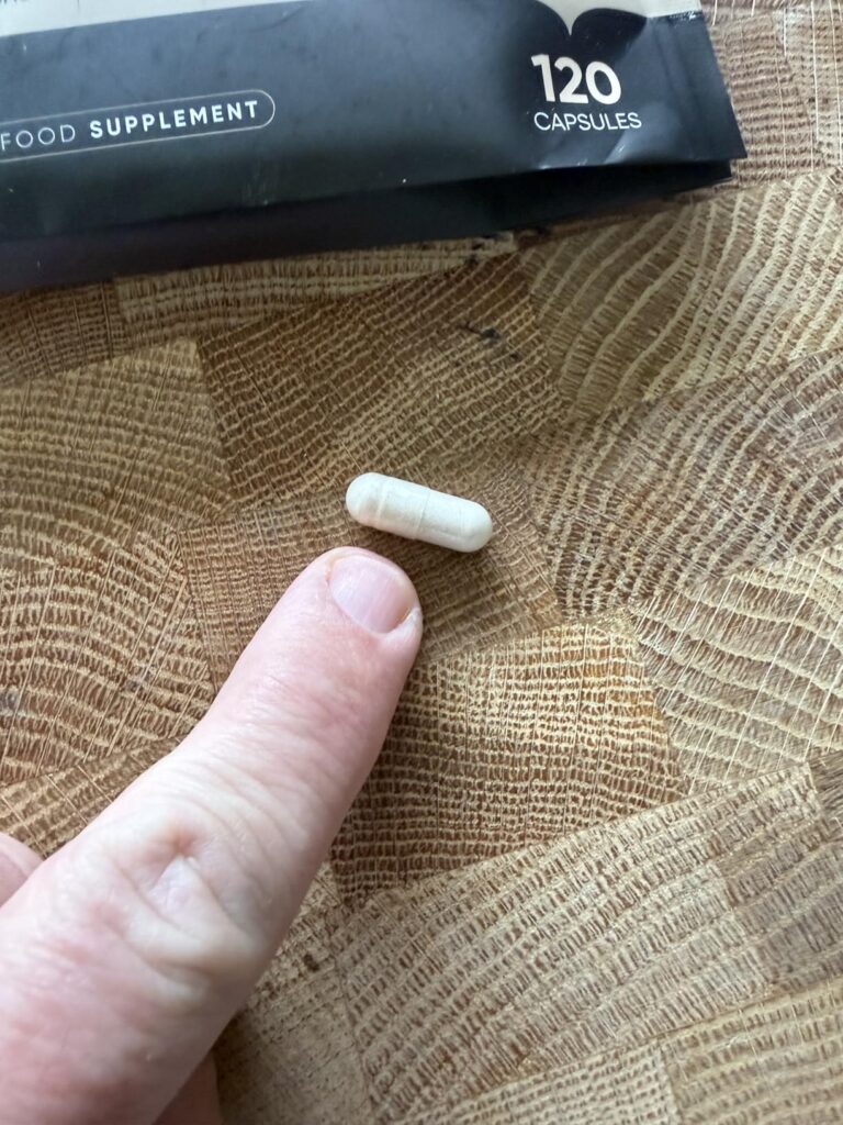 Une capsule de complément alimentaire est posée sur une planche en bois, avec un doigt qui la pointe.