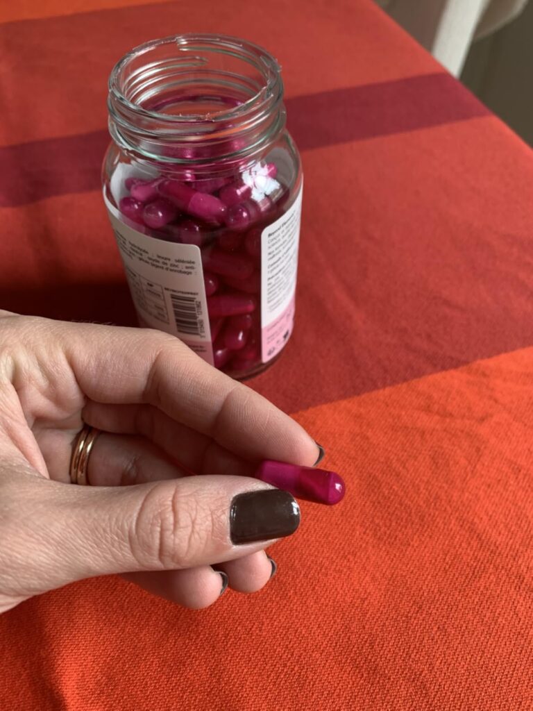 Une main tenant une gélule rose, le pot de gélules en arrière-plan sur une table rouge.
