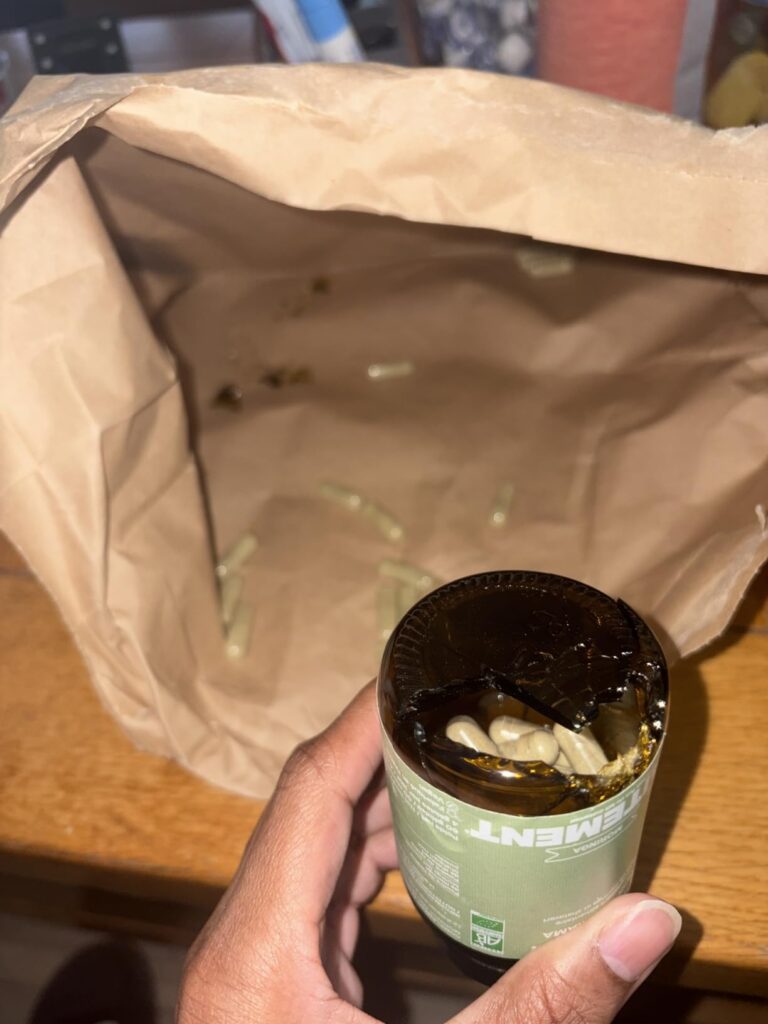 Une main tient un flacon de compléments alimentaires, visiblement abîmé, au-dessus d'un sac en papier kraft ouvert et contena...