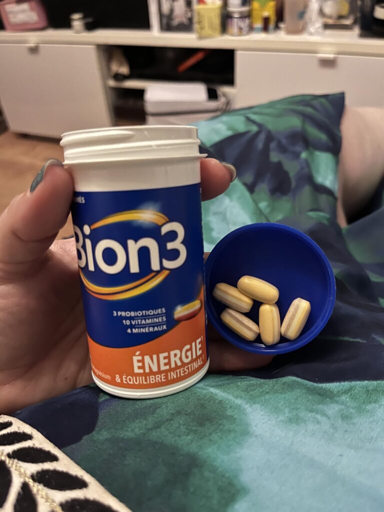 Une main tient le flacon de Bion3, à côté d'une coupelle avec des comprimés, sur un fond de tissu et d'intérieur.