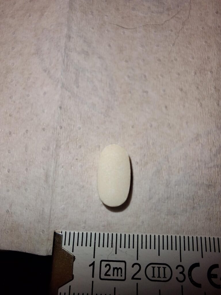 Une pilule blanche, en forme de gélule, posée à la verticale sur une surface blanche, avec une règle de mesure en dessous. Vu...