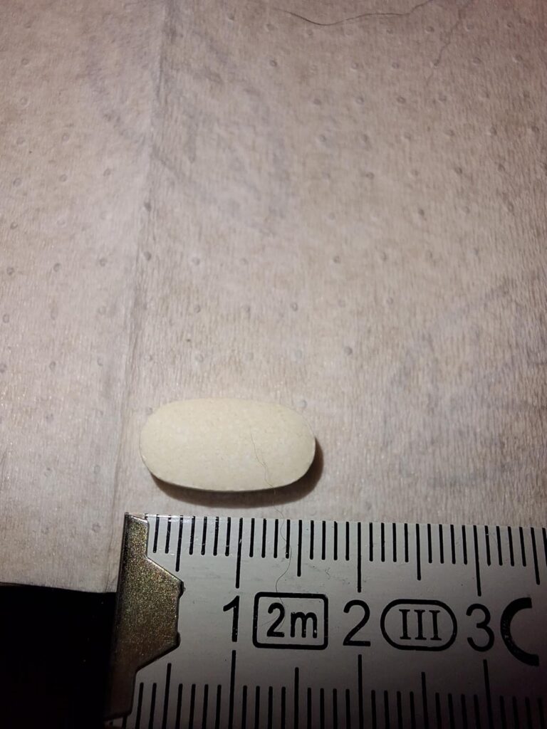 Une pilule blanche, en forme de gélule, posée à l'horizontale sur une surface blanche, avec une règle de mesure en dessous. V...